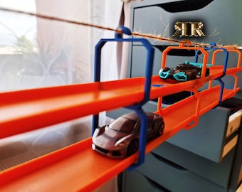 Könnte beinhalten: Ein zweistöckiges orangefarbenes Gleisset für Spielzeugautos aus Kunststoff mit blauen Stützen. Die Strecke ist an einer Kommodenschublade aufgehängt und verfügt über zwei Spielzeugautos, ein silbernes und ein blau-schwarzes.
