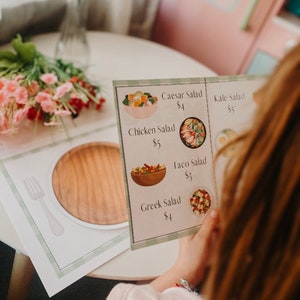 May include: A colourful menu with a green background lists different salad options with prices. The menu includes Caesar Salad, Kale Salad, Chicken Salad, Taco Salad, and Greek Salad. Each salad is illustrated with a picture of the salad in a bowl.