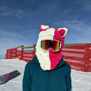 May include: A person wearing a white and pink crocheted ski mask with rainbow ski goggles. The person is wearing a teal ski suit and is standing on a snowy mountain.