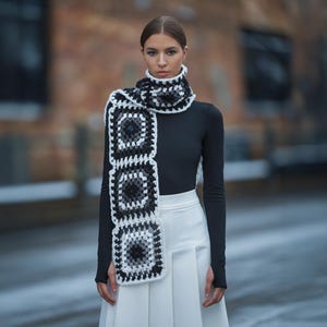 May include: A black and white crocheted scarf with a geometric pattern. The scarf is worn around the neck of a person wearing a black turtleneck and a white skirt.