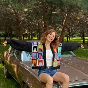 May include: A black, open-front cardigan with a colorful, square cat design. The cardigan is worn over a white top and blue denim shorts. The person is sitting on the hood of a brown car, with a green background of trees.