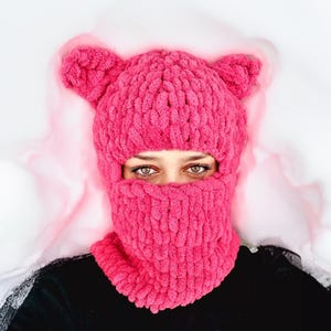 May include: A pink knitted balaclava with ear flaps. The balaclava is made of a thick, fuzzy yarn and covers the person's face, leaving only their eyes visible.