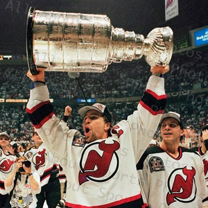 Könnte beinhalten: Eine Gruppe von Eishockeyspielern feiert mit der Stanley Cup Trophäe. Der Spieler in der Mitte hält die Trophäe über seinen Kopf und trägt ein weißes Trikot mit einem roten Teufel-Logo und der Nummer 20. Die anderen Spieler tragen ähnliche Trikots mit unterschiedlichen Nummern.