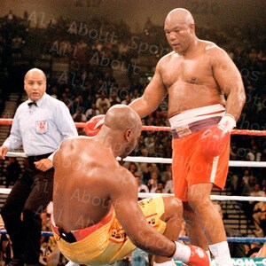 May include: A boxing match in a ring with two boxers. The boxer on the left is wearing yellow shorts with a red stripe and white boxing gloves. The boxer on the right is wearing red shorts with a white belt and white boxing gloves. The referee is standing in the background.