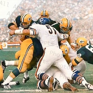 Puede incluir: Un grupo de jugadores de fútbol americano con uniformes verdes y blancos están en una concentración durante un partido. Los jugadores llevan cascos y protecciones. Los jugadores están en un campo verde con una multitud de personas en el fondo.