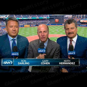 May include: Three men wearing blue suits and ties are sitting at a table. They are all smiling and looking at the camera. The man on the left is holding a microphone. The man in the middle is holding a microphone. The man on the right is holding a microphone. The table has a sign that says "SNY" on it. The sign also has the names of the men on it: Ron Darling, Gary Cohen, and Keith Hernandez.