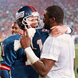 May include: Two football players in uniform embrace on the field after a game. The player on the left is wearing a blue jersey with the word "Giants" on it and the number "1". The player on the right is wearing a white jersey.