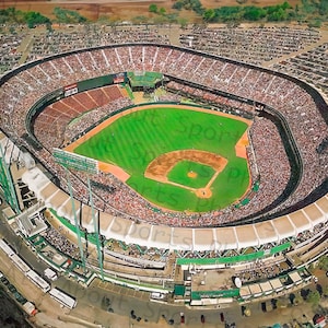 May include: An aerial view of a large, packed baseball stadium with a green field and white lines. The stadium is surrounded by a parking lot and trees.
