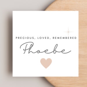 May include: A white square card with the words "PRECIOUS, LOVED, REMEMBERED" in black, and the name "Phoebe" in gray script. A light peach heart and a starburst graphic are also on the card. The card is on a light wood surface.