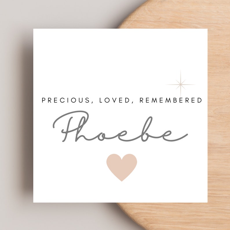 May include: A white square card with the words "PRECIOUS, LOVED, REMEMBERED" in black, and the name "Phoebe" in gray script. A light peach heart and a starburst graphic are also on the card. The card is on a light wood surface.