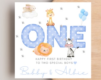 Op de afbeelding: Een vierkante verjaardagskaart met het woord ONE in grote blauwe letters met stippen. Illustraties bevatten een konijn op een wolk, een giraf, een leeuw en een zebra. Tekst: HAPPY FIRST BIRTHDAY TO TWO SPECIAL BOYS Bobby & Albie.
