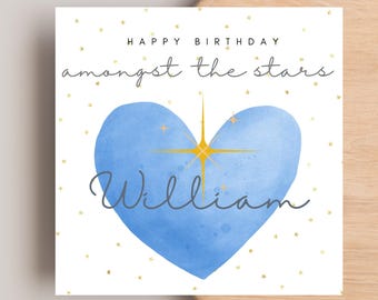 Op de afbeelding: Verjaardagskaart met een blauw aquarelhart en een gouden sterrenburst-ontwerp. De kaart leest "Happy Birthday" en "amongst the stars" met de naam "William" in script. Kleine gouden sterren verspreid over de witte achtergrond.