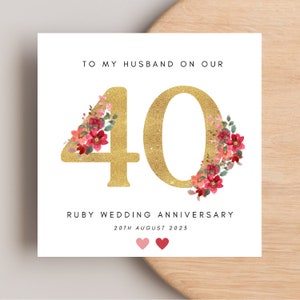 May include: A white card with gold glitter and red floral accents celebrating a 40th wedding anniversary. The text reads "To my husband on our 40th Ruby Wedding Anniversary 20th August 2023".