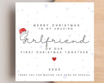 May include: A square greeting card with a white background featuring light blue snowflakes. The text reads: Merry Christmas to my amazing girlfriend on our first Christmas together, 2025, thank you for making this year so special. A red Santa hat and a small red heart are included.