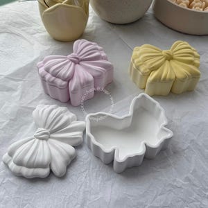 Puede incluir: Una colección de recipientes decorativos de cerámica en varias formas y colores. Incluye una caja en forma de lazo rosa claro, una caja en forma de lazo amarillo, un adorno en forma de lazo blanco y un plato en forma de hoja blanca.