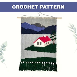 Puede incluir: Colgante de pared de crochet que representa una escena de paisaje con una casa, montañas y un árbol. La casa tiene un techo rojo y paredes blancas. Las montañas son azules y moradas. El árbol es verde. El colgante de pared tiene una franja verde.