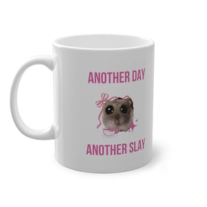 Könnte beinhalten: Weiße Keramiktasse mit dem Text "ANOTHER DAY ANOTHER SLAY" in Rosa. Die Tasse zeigt eine Grafik eines Hamsters mit einer rosa Schleife und einer Teetasse. Die Tasse hat einen gebogenen Henkel.