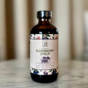 May include: A dark amber glass bottle of organic elderberry syrup. The label features the text "Organic Elderberry Syrup" and "Build Health + Combat Sickness". The bottle contains 8 fl oz (237mL) of syrup and is handcrafted in Ohio.