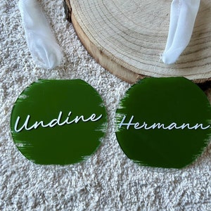 Könnte beinhalten: Zwei runde grüne Acryl-Ornamente mit weißen Pinselstrichen und weißer Schrift. Die Ornamente sagen "Undine" und "Hermann".