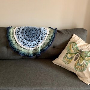 May include: A blue, green, and white crocheted throw blanket with a circular design and a fringe border. The blanket is draped over a gray couch with a white pillow featuring a green butterfly.