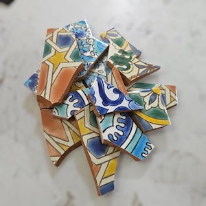 May include: A pile of broken ceramic tile fragments with detailed patterns. The pieces feature a mix of colours, such as blue, yellow, orange, and green, with geometric and floral designs. The tiles seem to be from a decorative project.