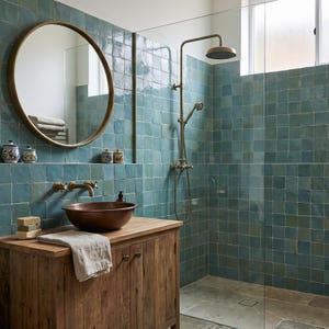 May include: A bathroom with teal square tiles, a wooden vanity with a copper sink, and a round gold mirror. The shower has a glass enclosure and a vintage-style showerhead and faucet. The room has a rustic, spa-like aesthetic.
