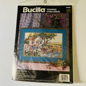 May include: A Bucilla counted cross-stitch kit featuring a colorful scene of a white cottage with a bridge and flowers. The kit includes 14 count Aida cloth, cotton embroidery floss, a needle, and easy-to-follow instructions with a chart. The kit is designed by Erin Dertner and measures 10 inches by 10 inches.