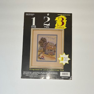 May include: A counted cross stitch kit featuring a charming cottage with a wishing well, flowers, and a path. The kit is part of a progressive level learning series, labeled "1, 2, 3" with the corresponding words "One, Two, Three". The kit is labeled "01242 Wishing Well" and includes instructions to become an expert.