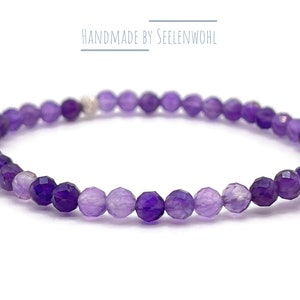 Könnte beinhalten: Ein zartes Armband mit facettierten lila Perlen in verschiedenen Schattierungen, von tiefem Violett bis Lavendel. Das Armband hat eine kleine silberne Akzentperle. Der Text "HANDMADE BY SEELENWOHL" befindet sich über dem Armband.