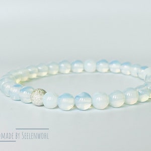 Könnte beinhalten: Ein zartes Armband aus hellblauen Opalperlen und einer einzelnen silbernen Perle. Das Armband ist handgefertigt und trägt den Text "HANDMADE BY SEELENWOHL" am unteren Rand des Bildes.