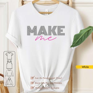 May include: White t-shirt featuring the text "MAKE me". The word "MAKE" is in black, striped letters, and "me" is in pink script. The shirt has a round neck and short sleeves. The shirt is displayed on a wooden hanger.