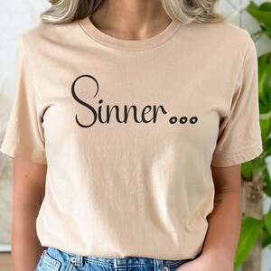 Könnte beinhalten: Ein beiges T-Shirt mit dem Aufdruck "Sinner..." in Schwarz.