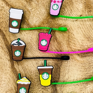 Puede incluir: Una colección de coloridos dijes de tazas de café con correas verdes, rosas y negras. Los dijes presentan el logotipo de Starbucks y varios diseños, incluyendo crema batida y tazas de diferentes colores.