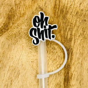 May include: A clear plastic straw with a white plastic accent and a white and black topper that reads "oh shit" in a cursive font. The straw is on a wooden surface.
