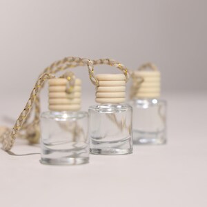 May include: Three small, clear glass bottles with wooden caps and a braided rope for hanging. The bottles are filled with a clear liquid. The rope is beige with gold accents. The bottles are arranged in a row.