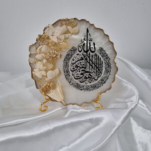 May include: White resin art piece with a gold trim and dried white flowers. The art piece features black Arabic calligraphy that reads "Allah" and a circle of smaller Arabic text. The art piece is on a gold stand.
