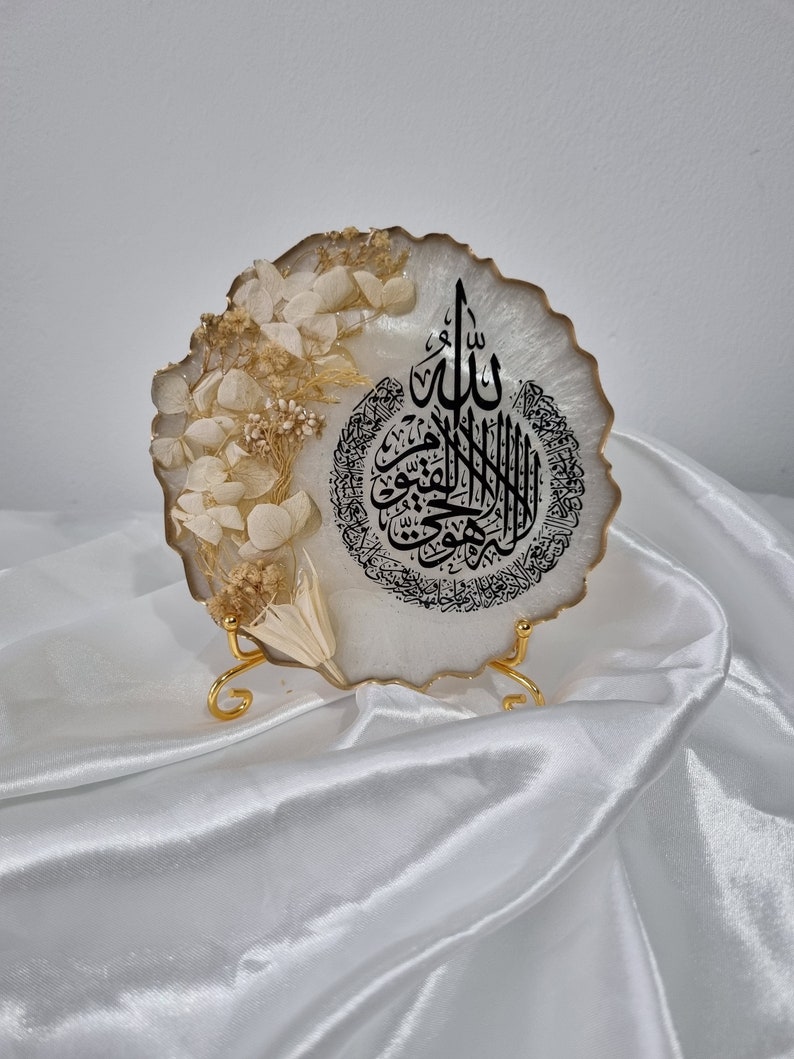 May include: White resin art piece with a gold trim and dried white flowers. The art piece features black Arabic calligraphy that reads "Allah" and a circle of smaller Arabic text. The art piece is on a gold stand.