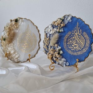 May include: Two round resin art pieces with gold calligraphy and dried flowers. The pieces are on gold metal stands and are displayed on a white surface.