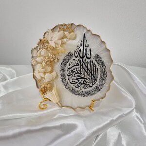 May include: A white resin round plaque with a gold rim and a black Arabic calligraphy design. The plaque is decorated with dried white flowers and is displayed on a gold metal stand.