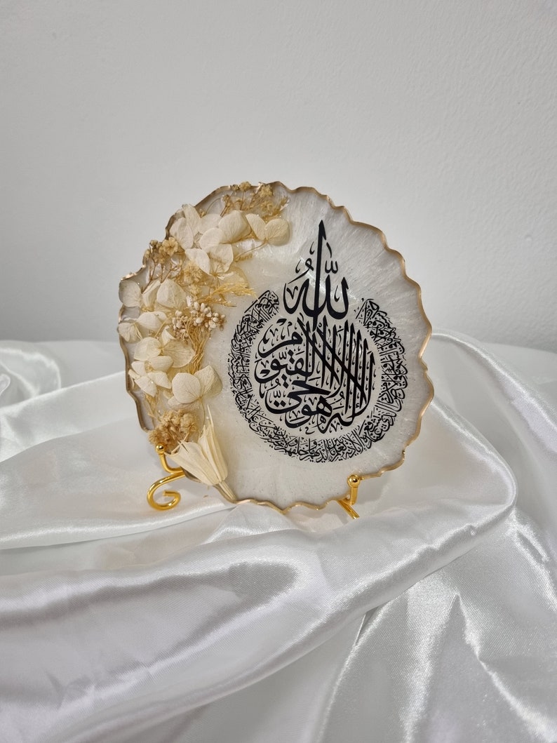 May include: A white resin round plaque with a gold rim and a black Arabic calligraphy design. The plaque is decorated with dried white flowers and is displayed on a gold metal stand.