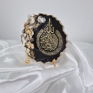 May include: A black resin wall decor with gold calligraphy and dried white flowers. The decor is on a gold stand and has a scalloped edge.