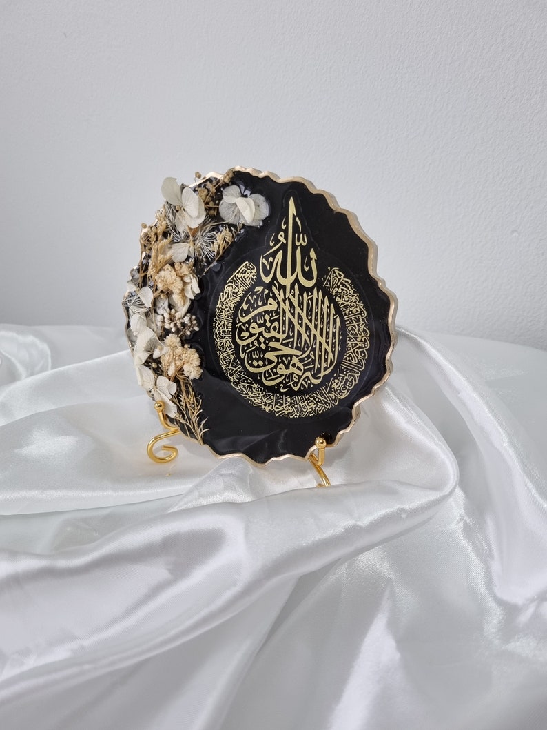 May include: A black resin wall decor with gold calligraphy and dried white flowers. The decor is on a gold stand and has a scalloped edge.