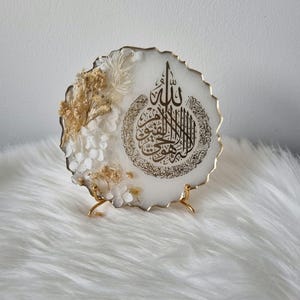 May include: White resin round plaque with a gold rim and a gold Arabic calligraphy design. The plaque is decorated with dried white and beige flowers and is displayed on a gold metal stand.