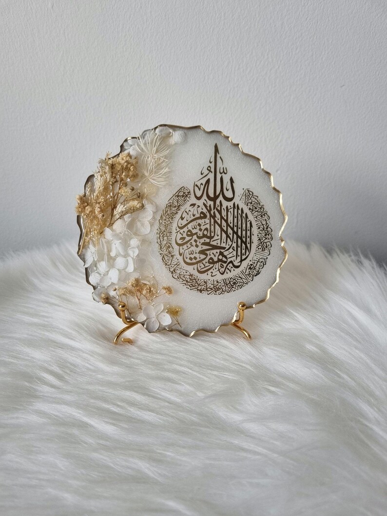 May include: White resin round plaque with a gold rim and a gold Arabic calligraphy design. The plaque is decorated with dried white and beige flowers and is displayed on a gold metal stand.