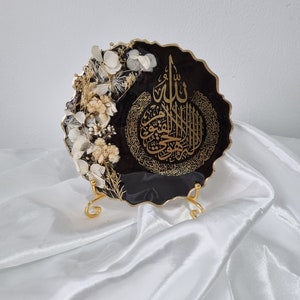 May include: A black resin art piece with a gold metallic finish. The piece features a decorative, intricate Arabic calligraphy design and dried white flowers. The art piece is displayed on a gold metal stand.