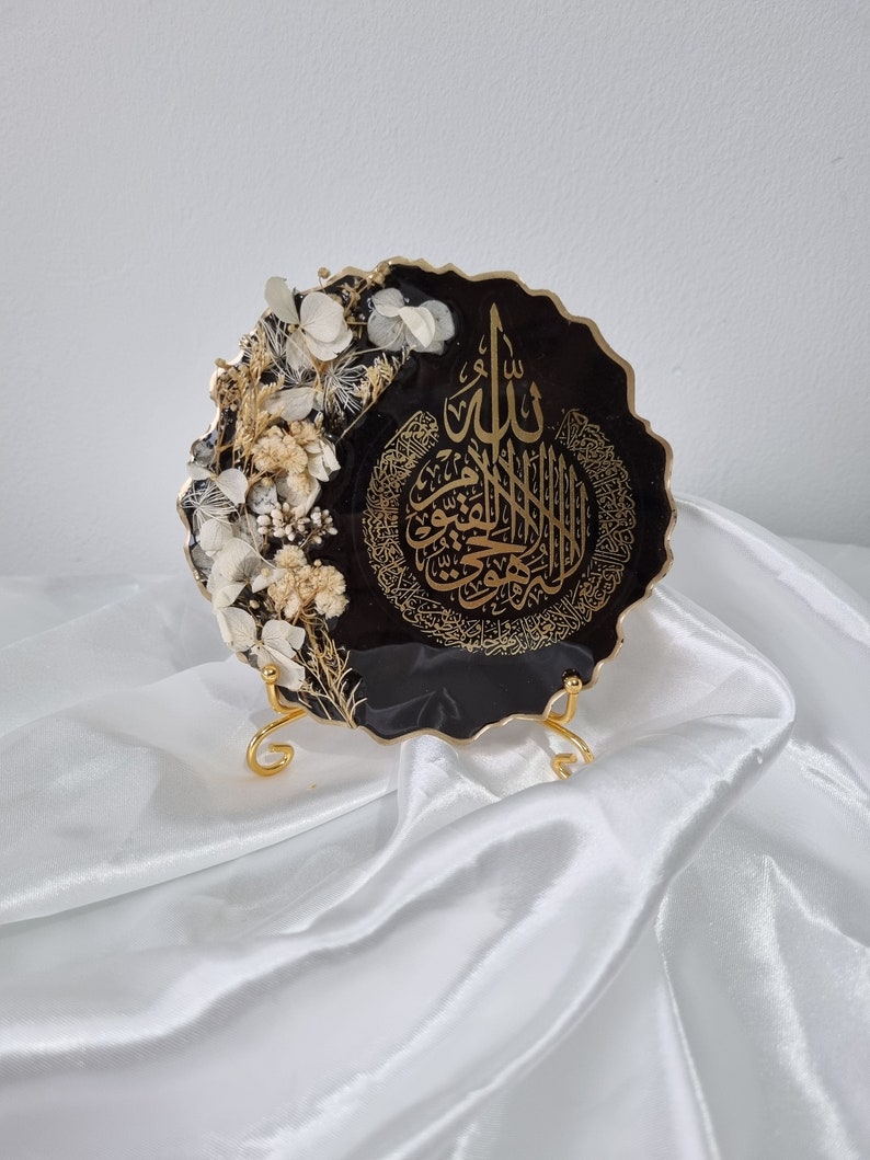 May include: A black resin art piece with a gold metallic finish. The piece features a decorative, intricate Arabic calligraphy design and dried white flowers. The art piece is displayed on a gold metal stand.