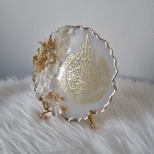 May include: White resin round tray with a gold metallic finish. The tray features a gold Arabic calligraphy design and dried flowers. The tray is on a gold metal stand.