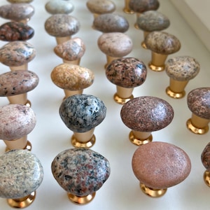 May include: A collection of natural stone drawer pulls with gold metal bases. The stones are various shades of brown, gray, and pink.