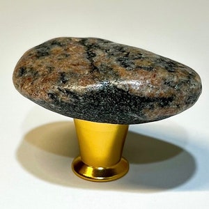 Könnte beinhalten: Ein natürlicher Steinknauf für Schränke mit einem braunen, schwarzen und rosafarbenen, gesprenkelten Muster. Der Knauf ist an einer goldenen Metallbasis befestigt.