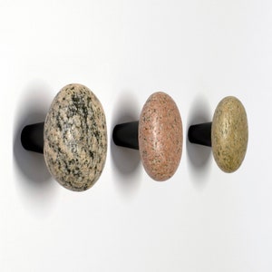 Könnte beinhalten: Drei Steinhaken in verschiedenen Farben und Größen, an einer weißen Wand montiert. Die Haken sind in Form von abgerundeten Steinen und haben schwarze Metallsockel.